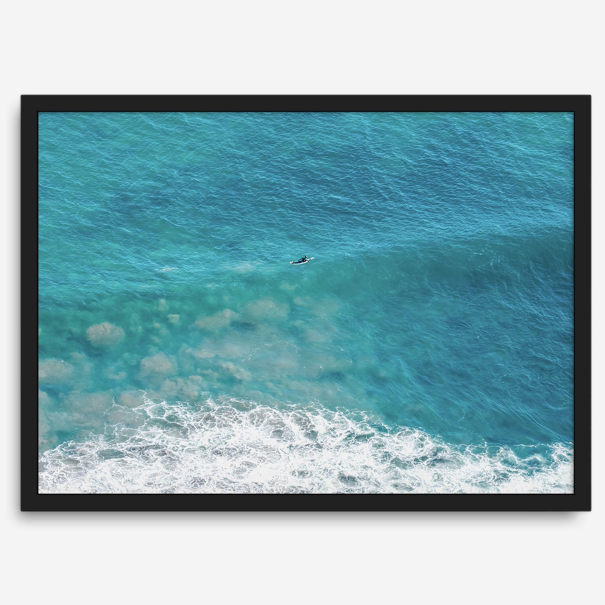 Aerial beach wall art featuring a lone surfer paddling through turquoise ocean waves, with foamy white surf along the shoreline.