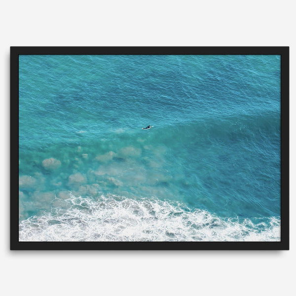 Aerial beach wall art featuring a lone surfer paddling through turquoise ocean waves, with foamy white surf along the shoreline.
