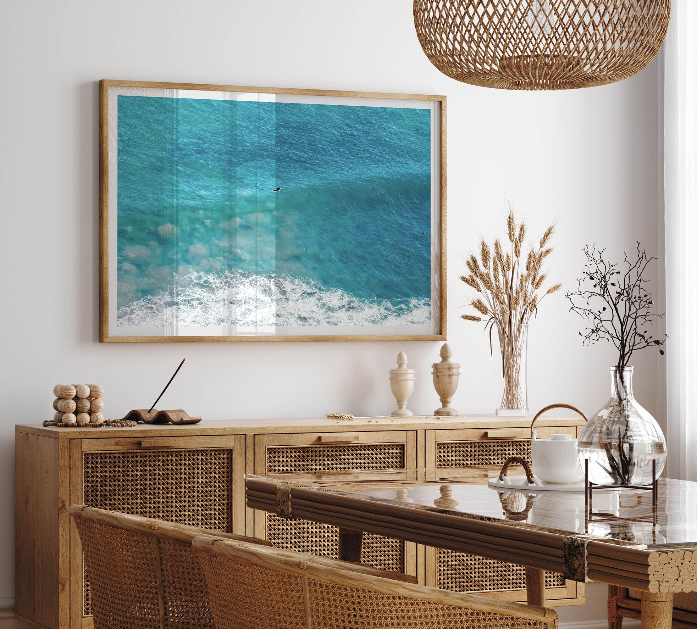 Aerial beach wall art featuring a lone surfer paddling through turquoise ocean waves, with foamy white surf along the shoreline.