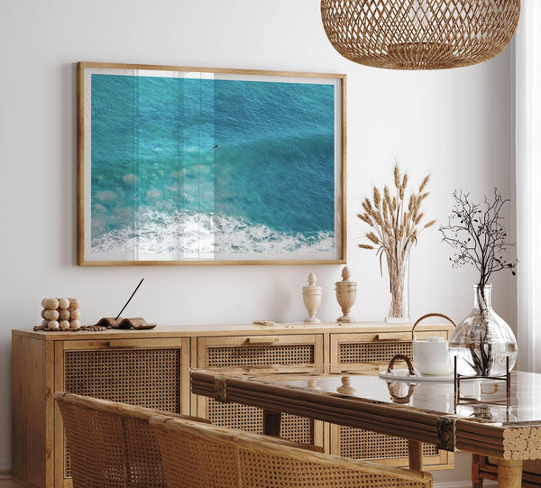 Aerial beach wall art featuring a lone surfer paddling through turquoise ocean waves, with foamy white surf along the shoreline.