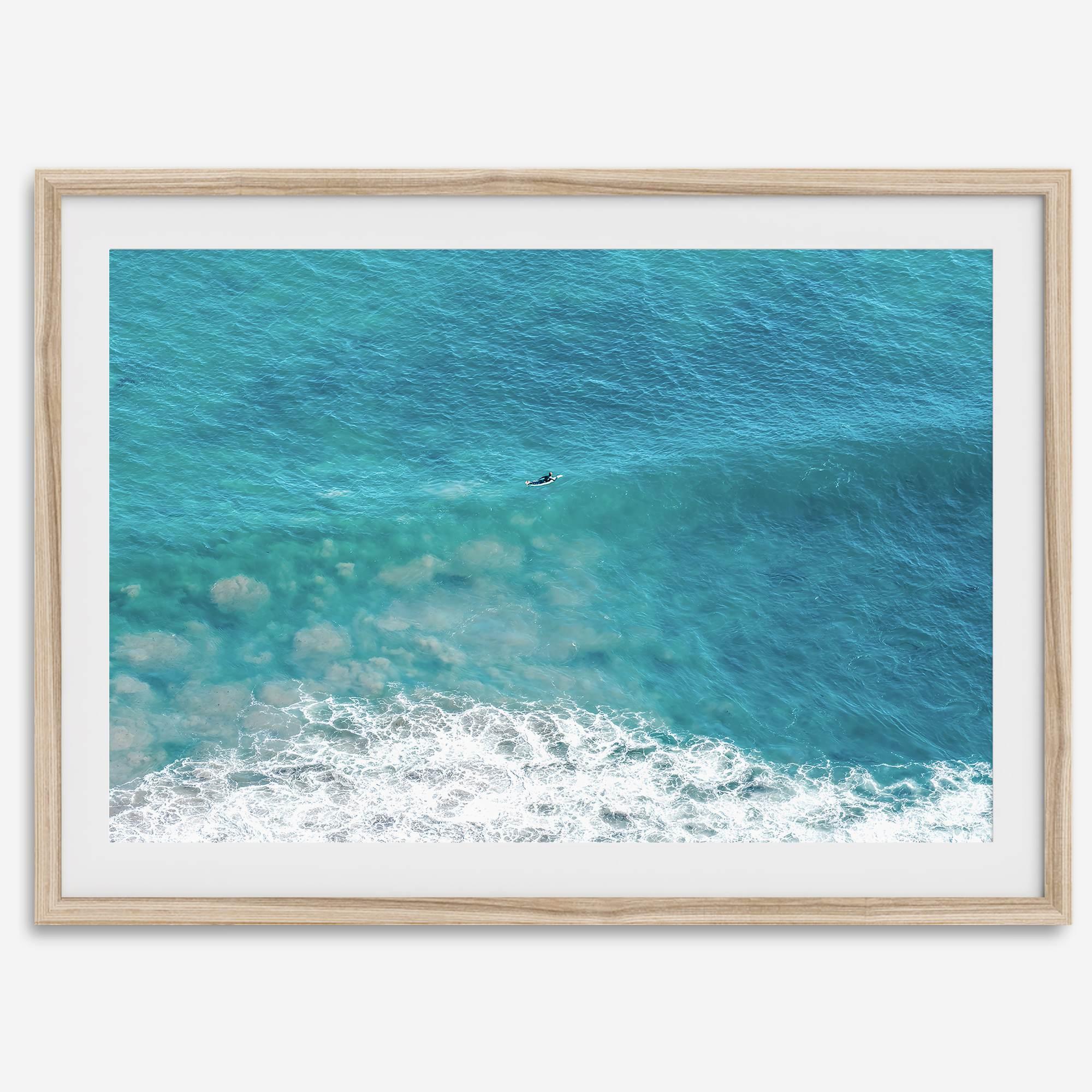 Aerial beach wall art featuring a lone surfer paddling through turquoise ocean waves, with foamy white surf along the shoreline.