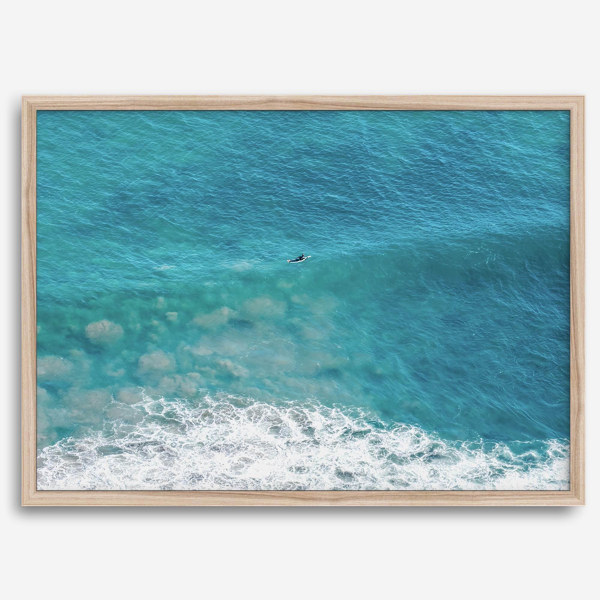 Aerial beach wall art featuring a lone surfer paddling through turquoise ocean waves, with foamy white surf along the shoreline.