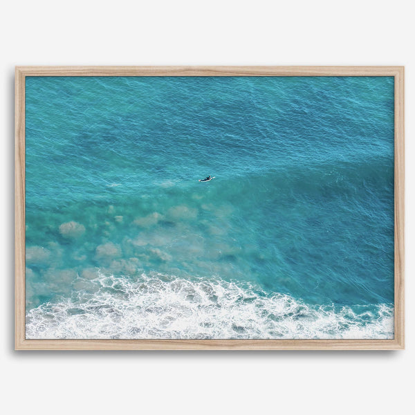 Aerial beach wall art featuring a lone surfer paddling through turquoise ocean waves, with foamy white surf along the shoreline.