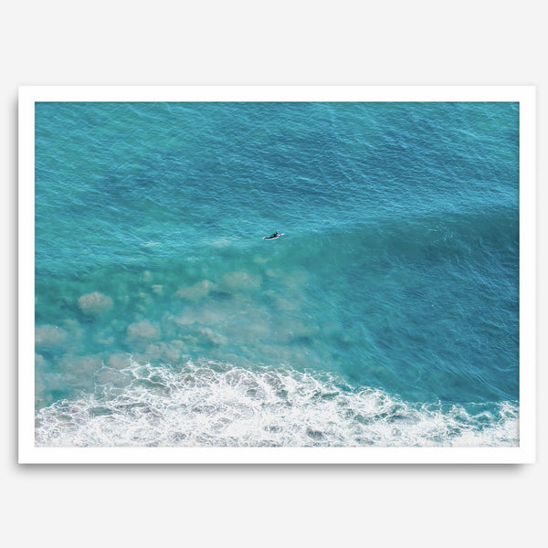 Aerial beach wall art featuring a lone surfer paddling through turquoise ocean waves, with foamy white surf along the shoreline.