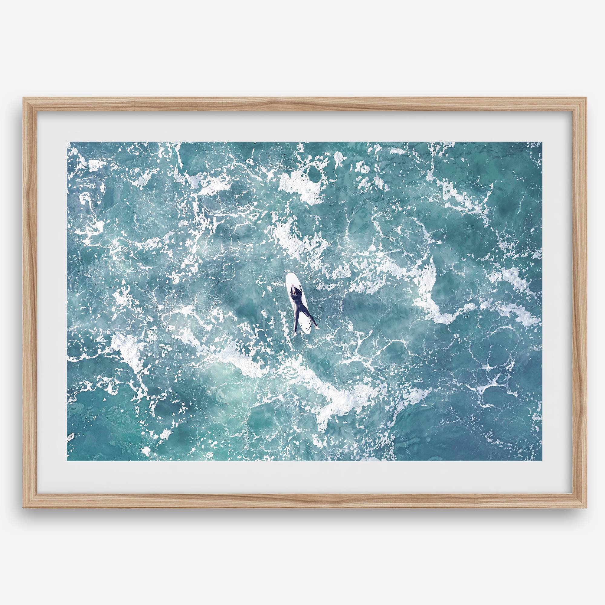 Fine art aerial photography print of a lone surfer floating on a surfboard amidst turquoise ocean waves with intricate white foam patterns.