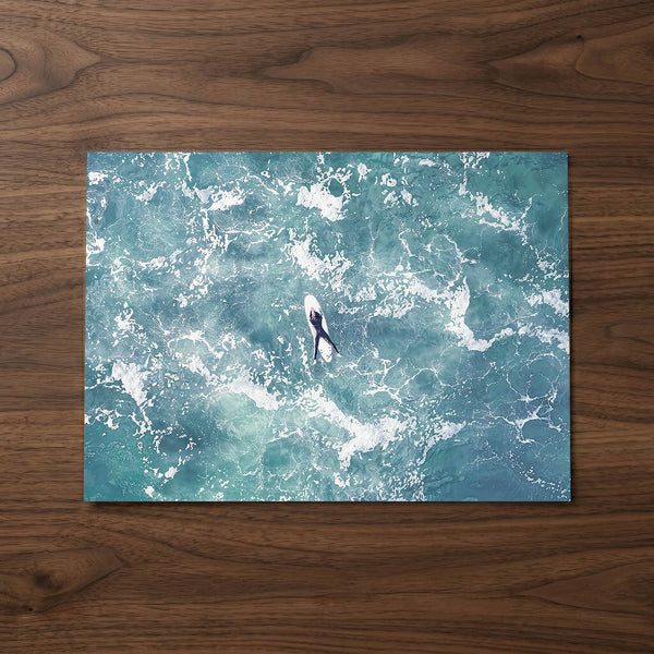 Fine art aerial photography print of a lone surfer floating on a surfboard amidst turquoise ocean waves with intricate white foam patterns.