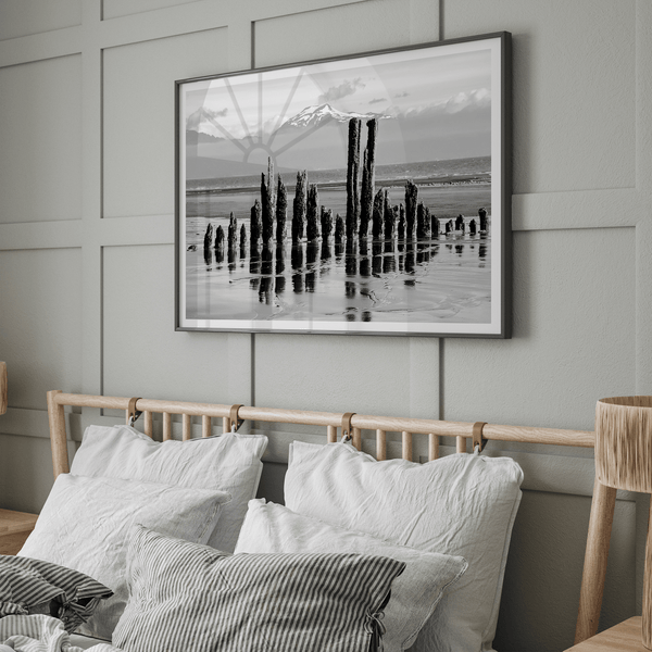 Black and white photo of weathered pilings on an Alaskan beach with snow-capped mountains in the background – fine art print.