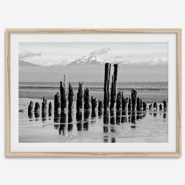 Black and white photo of weathered pilings on an Alaskan beach with snow-capped mountains in the background – fine art print.