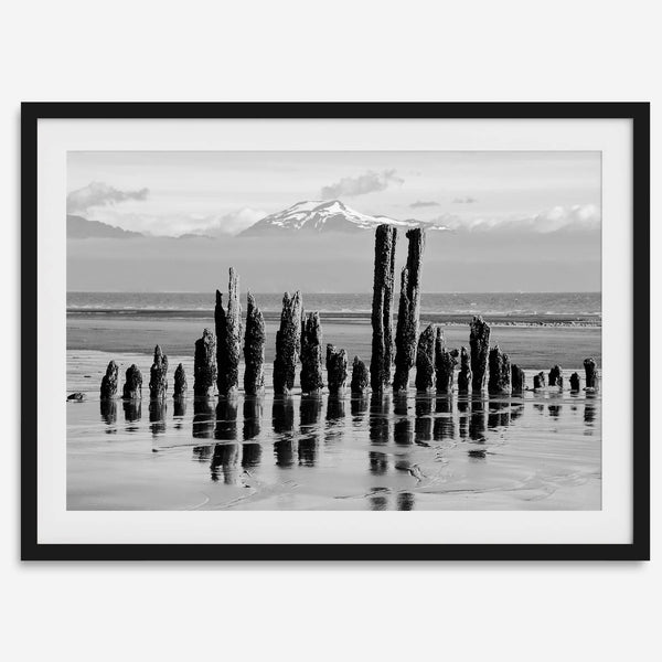 Black and white photo of weathered pilings on an Alaskan beach with snow-capped mountains in the background – fine art print.