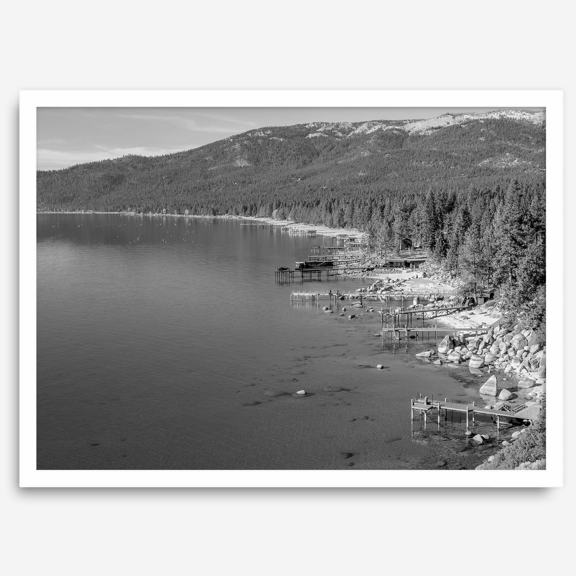 A fine art black and white Lake Tahoe print showcasing docks and boats nested on the lake waters on the left, while the beachfront, nestled against a backdrop of a lush forest and majestic mountain, paints a picturesque scene on the right.