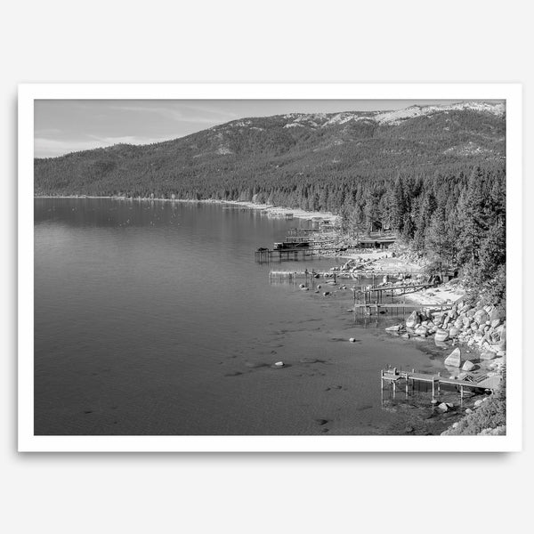 A fine art black and white Lake Tahoe print showcasing docks and boats nested on the lake waters on the left, while the beachfront, nestled against a backdrop of a lush forest and majestic mountain, paints a picturesque scene on the right.