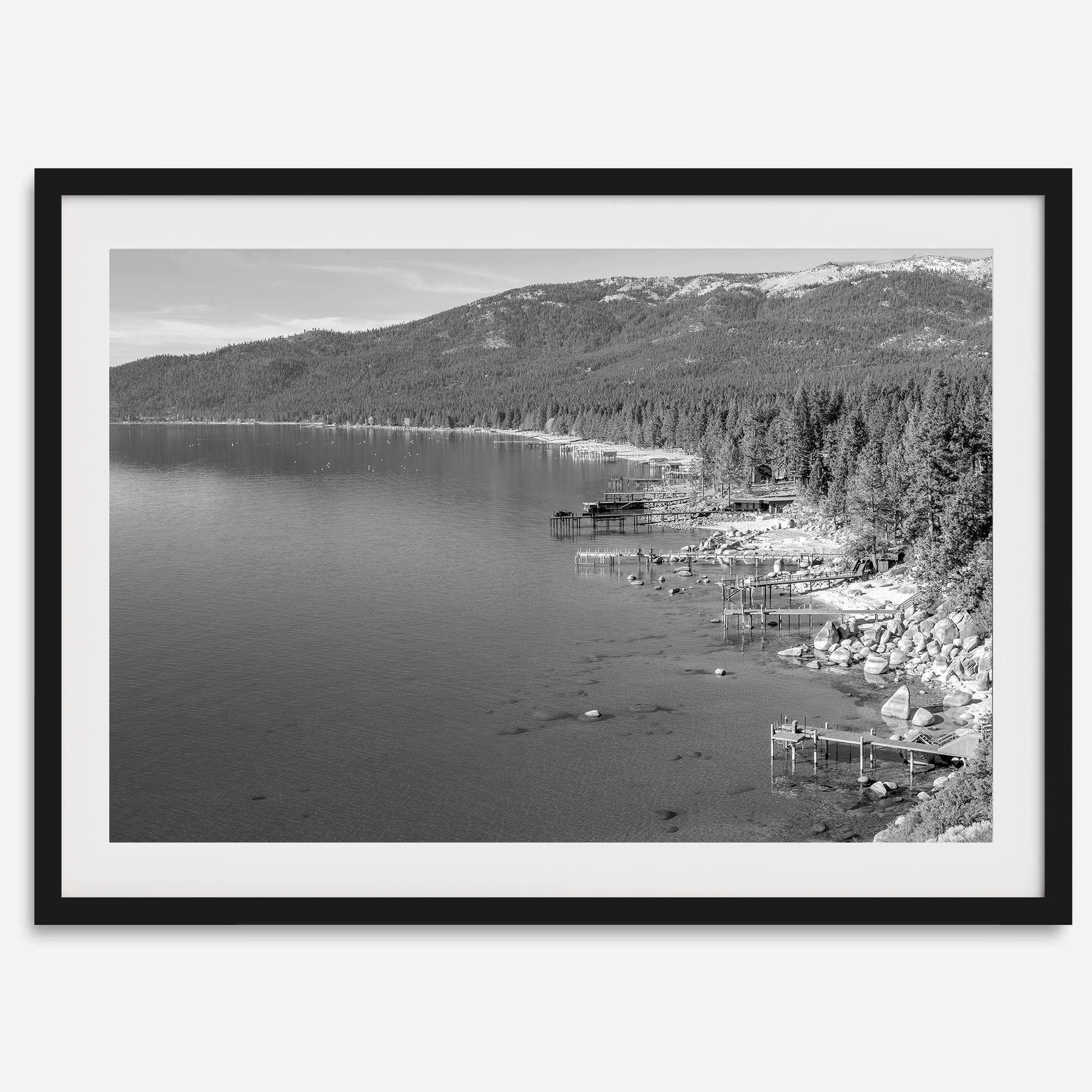 A fine art black and white Lake Tahoe print showcasing docks and boats nested on the lake waters on the left, while the beachfront, nestled against a backdrop of a lush forest and majestic mountain, paints a picturesque scene on the right.