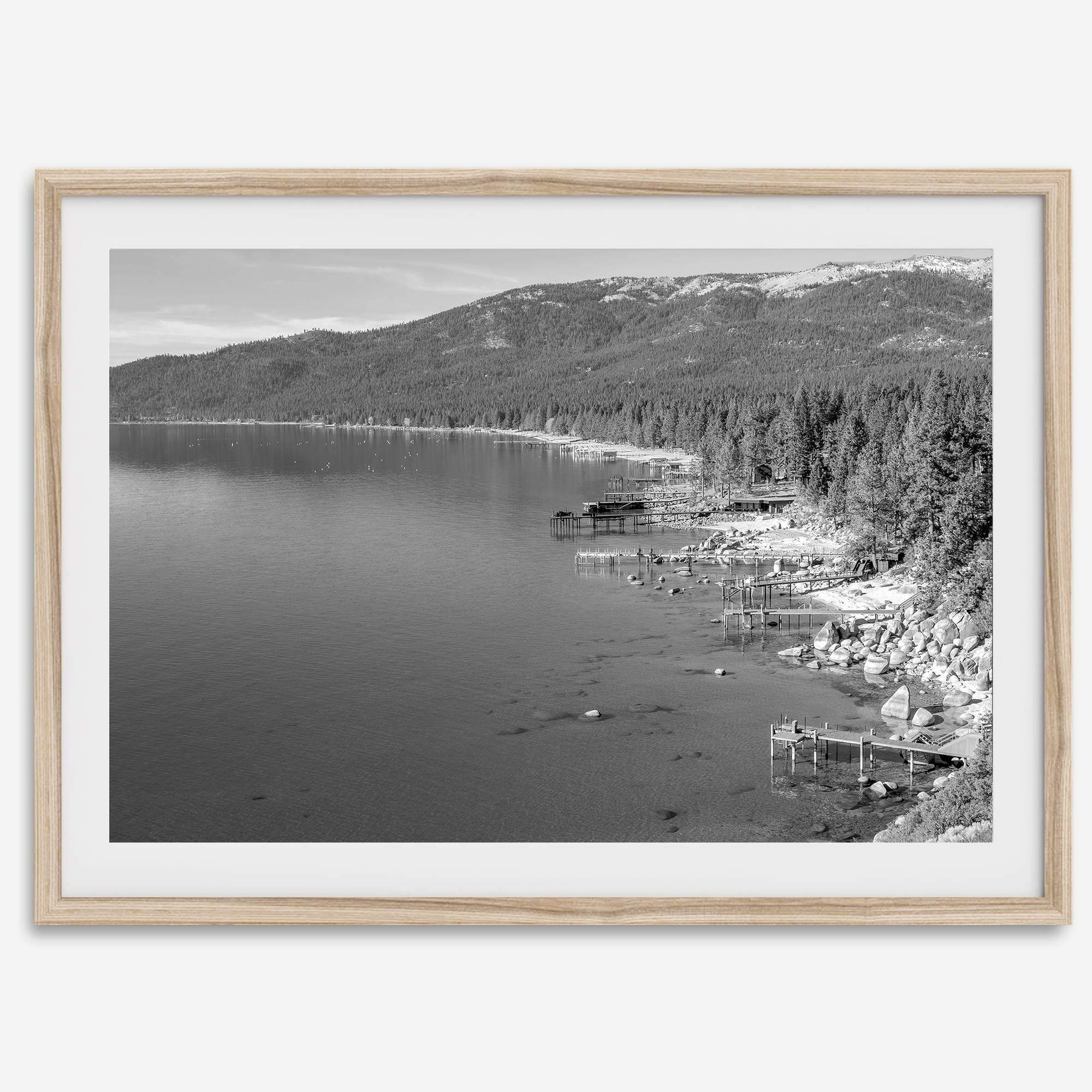 A fine art black and white Lake Tahoe print showcasing docks and boats nested on the lake waters on the left, while the beachfront, nestled against a backdrop of a lush forest and majestic mountain, paints a picturesque scene on the right.