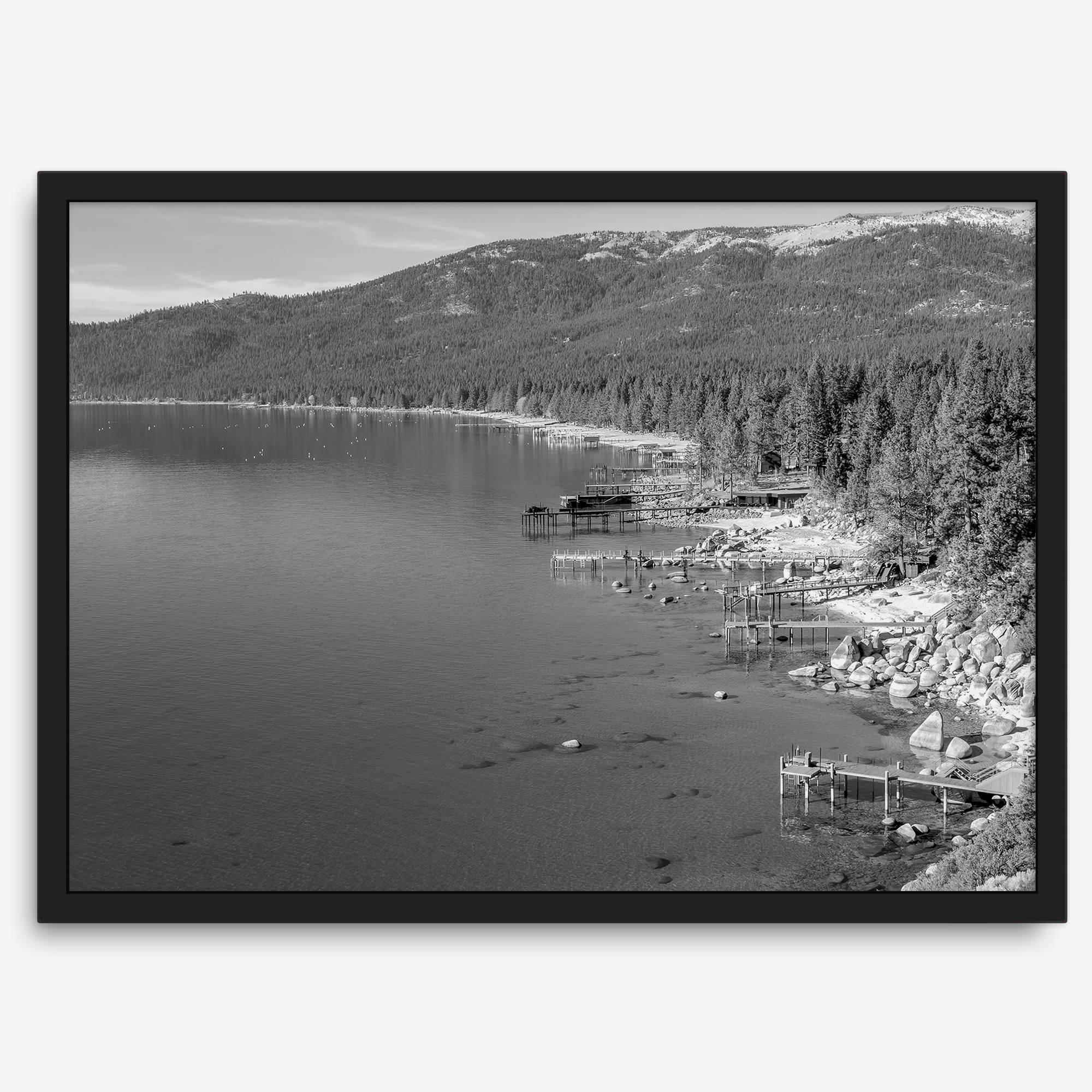 A fine art black and white Lake Tahoe print showcasing docks and boats nested on the lake waters on the left, while the beachfront, nestled against a backdrop of a lush forest and majestic mountain, paints a picturesque scene on the right.