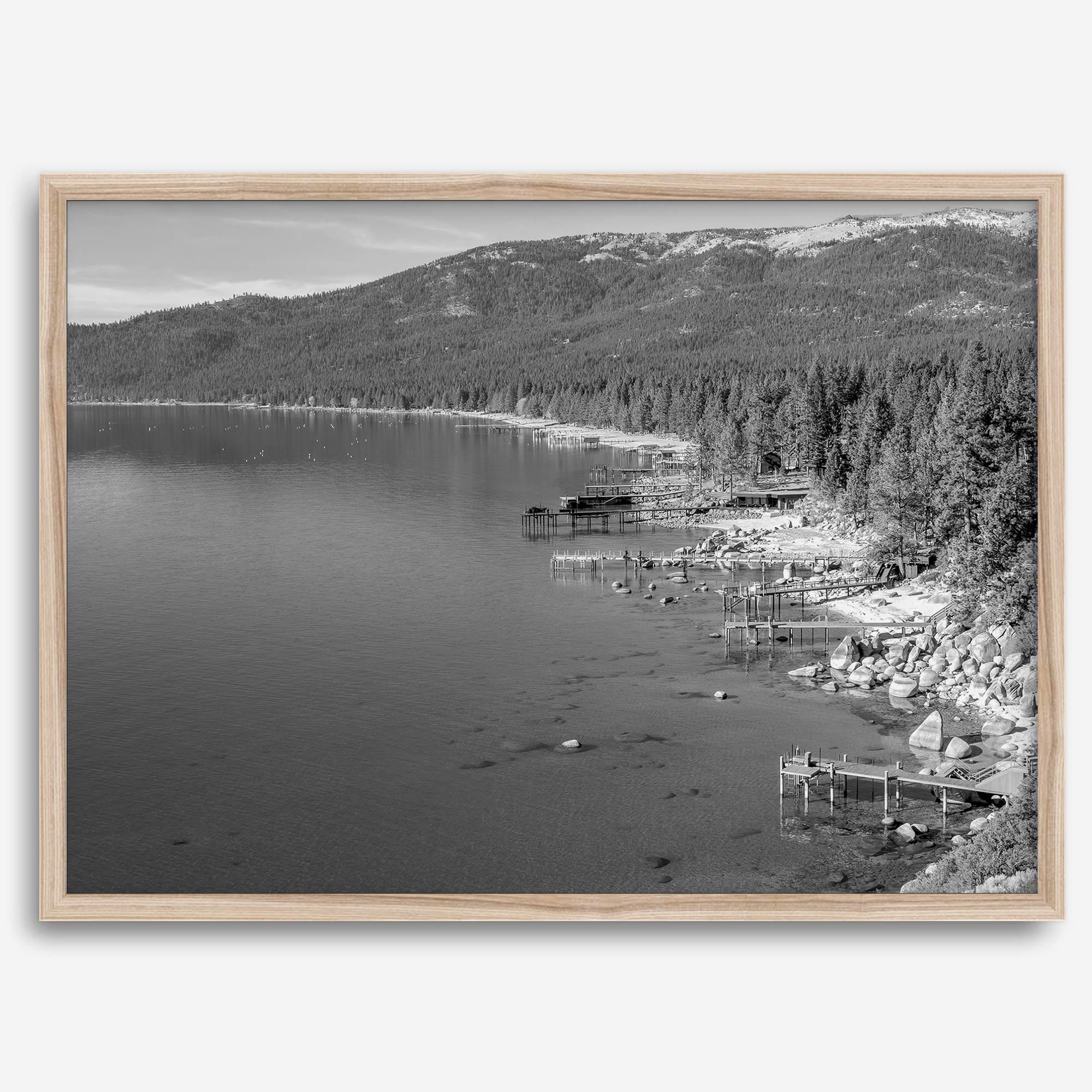 A fine art black and white Lake Tahoe print showcasing docks and boats nested on the lake waters on the left, while the beachfront, nestled against a backdrop of a lush forest and majestic mountain, paints a picturesque scene on the right.