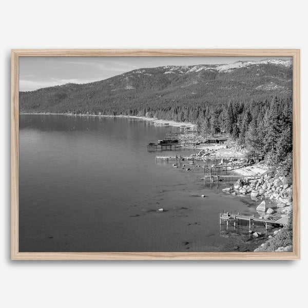 A fine art black and white Lake Tahoe print showcasing docks and boats nested on the lake waters on the left, while the beachfront, nestled against a backdrop of a lush forest and majestic mountain, paints a picturesque scene on the right.