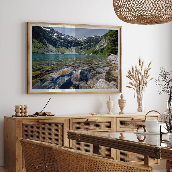 A framed fine art landscape print of a crystal-clear blue lake in Washington State. The tranquil water reveals colorful rocks beneath the surface, with snow-capped mountains towering in the distance.