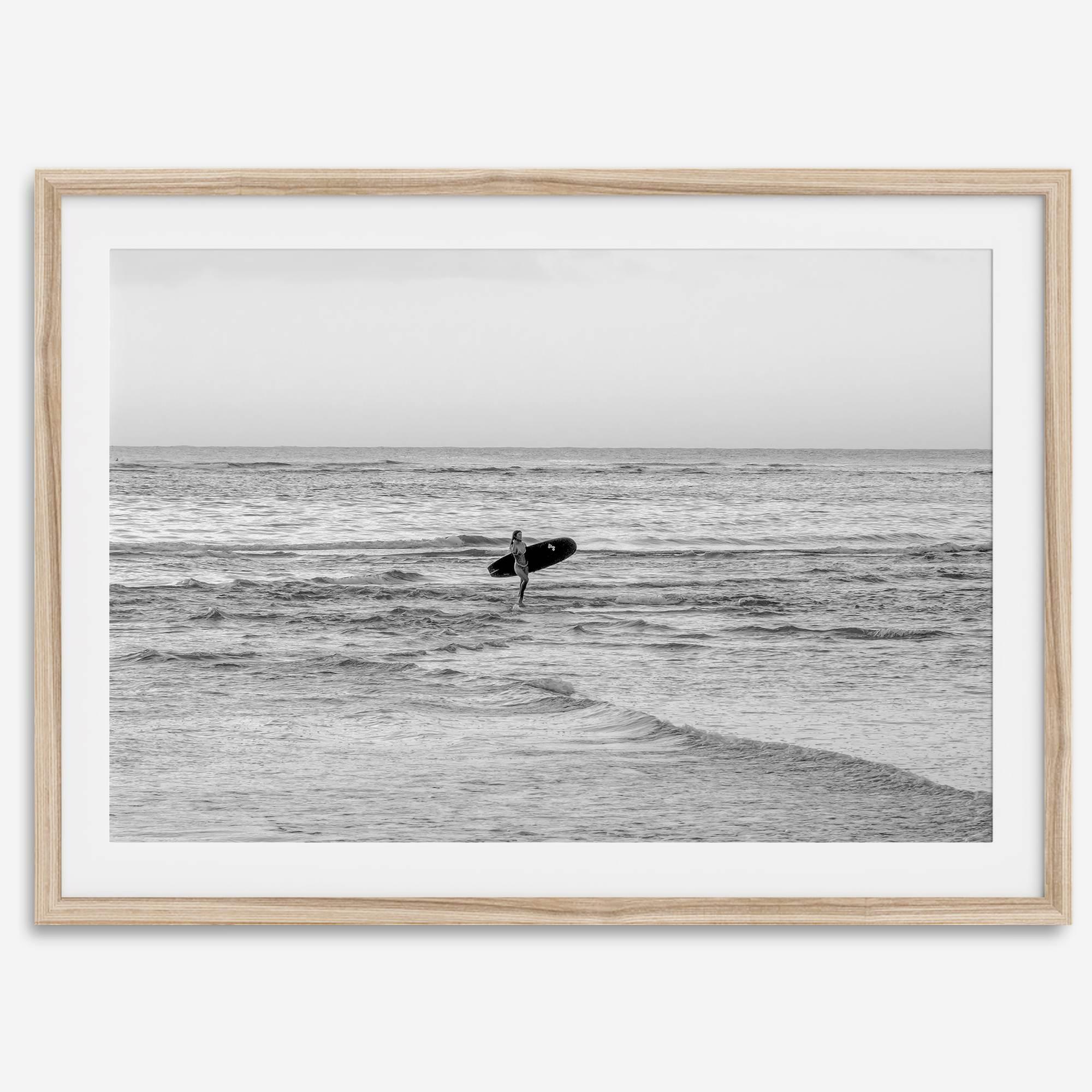 A fine art photography print capturing a female surfer walking back from the ocean at sunset with waves breaking in the foreground, capturing the end of a surf session.