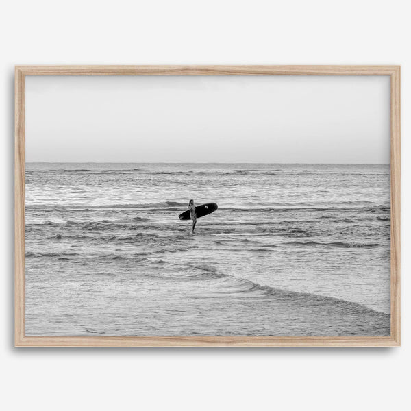 A fine art photography print capturing a female surfer walking back from the ocean at sunset with waves breaking in the foreground, capturing the end of a surf session.