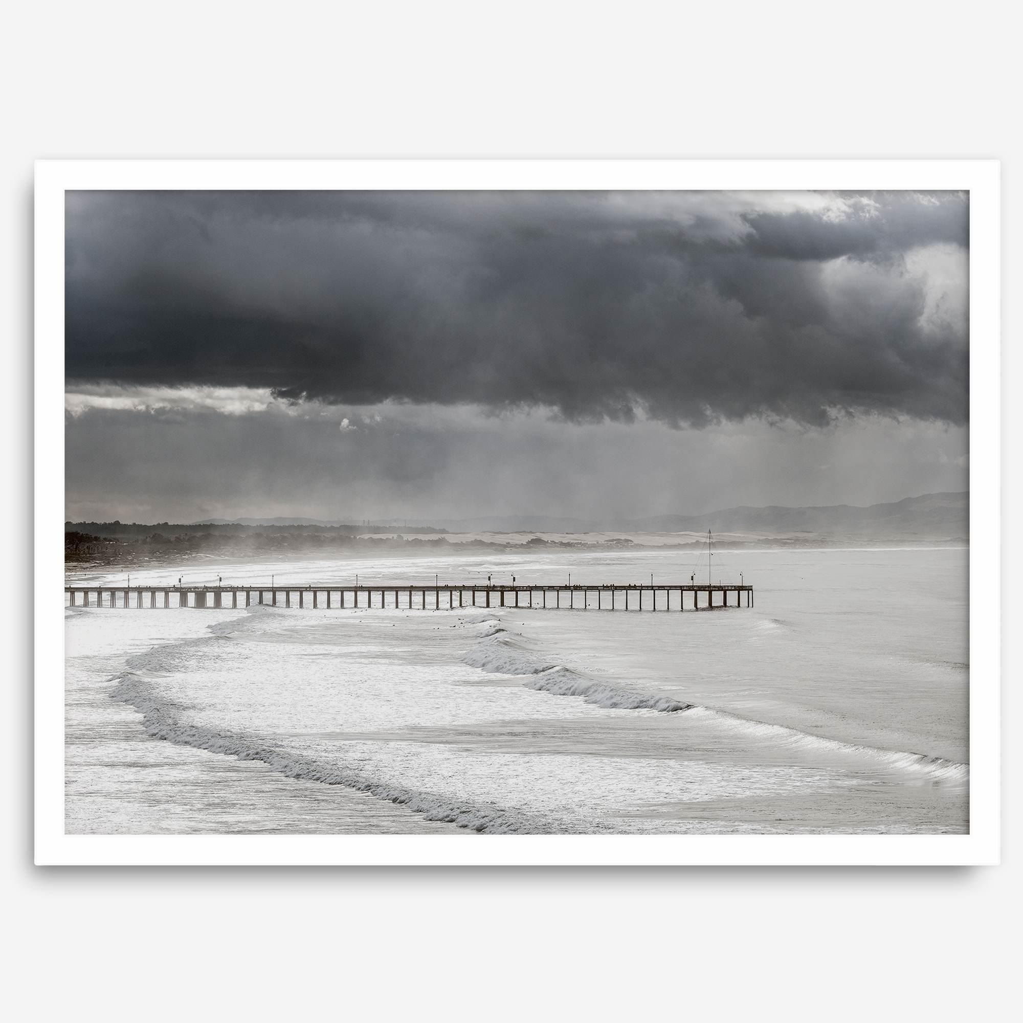 This fine art ocean photography print was taken in Pismo Beach, California, and showcases a distant view of an ocean pier and stormy clouds. Looking long and hard, you can see tens of surfers around the pier.