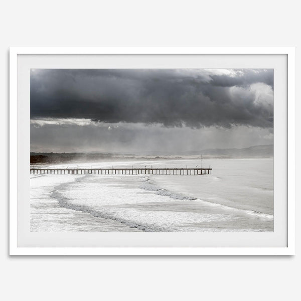 This fine art ocean photography print was taken in Pismo Beach, California, and showcases a distant view of an ocean pier and stormy clouds. Looking long and hard, you can see tens of surfers around the pier.