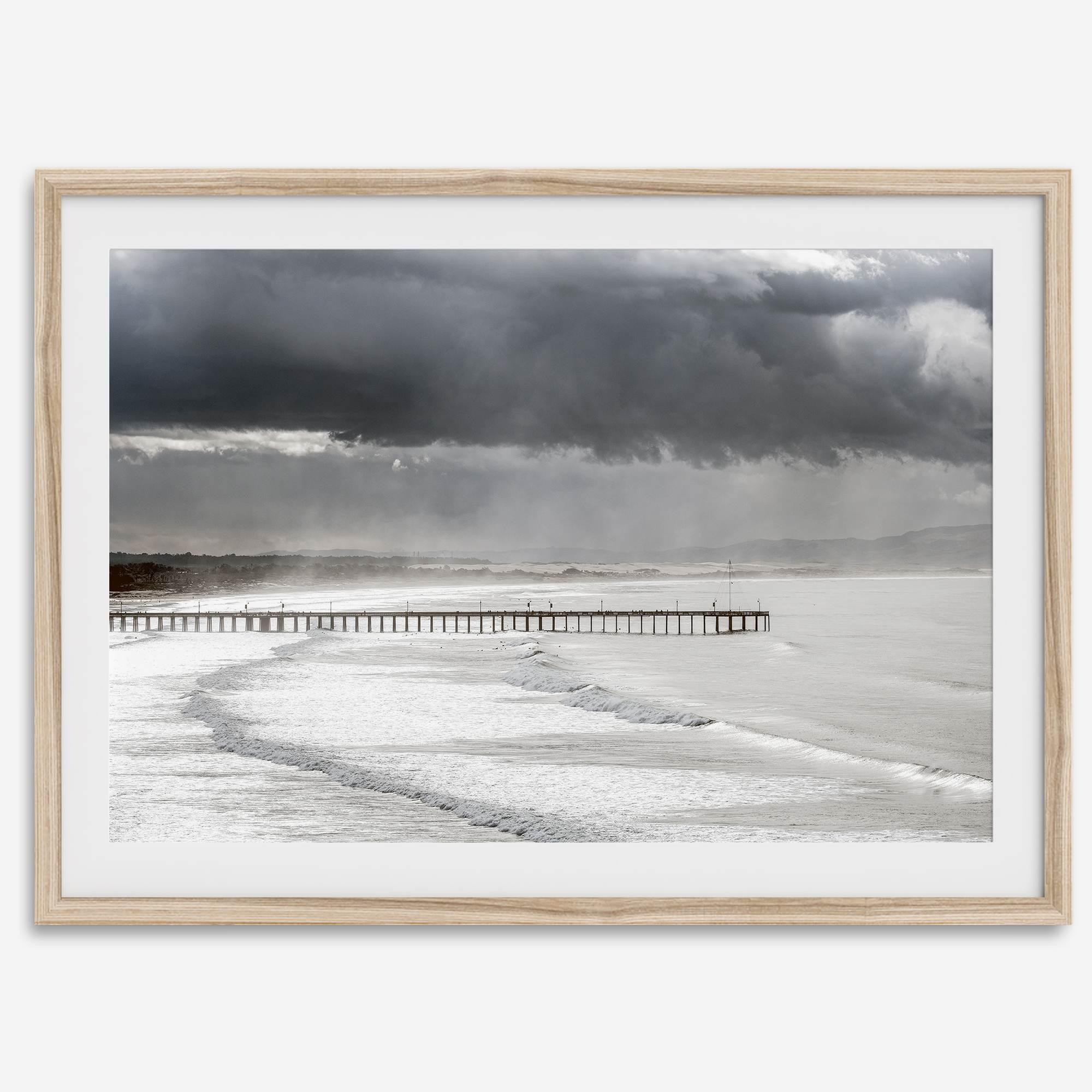 This fine art ocean photography print was taken in Pismo Beach, California, and showcases a distant view of an ocean pier and stormy clouds. Looking long and hard, you can see tens of surfers around the pier.