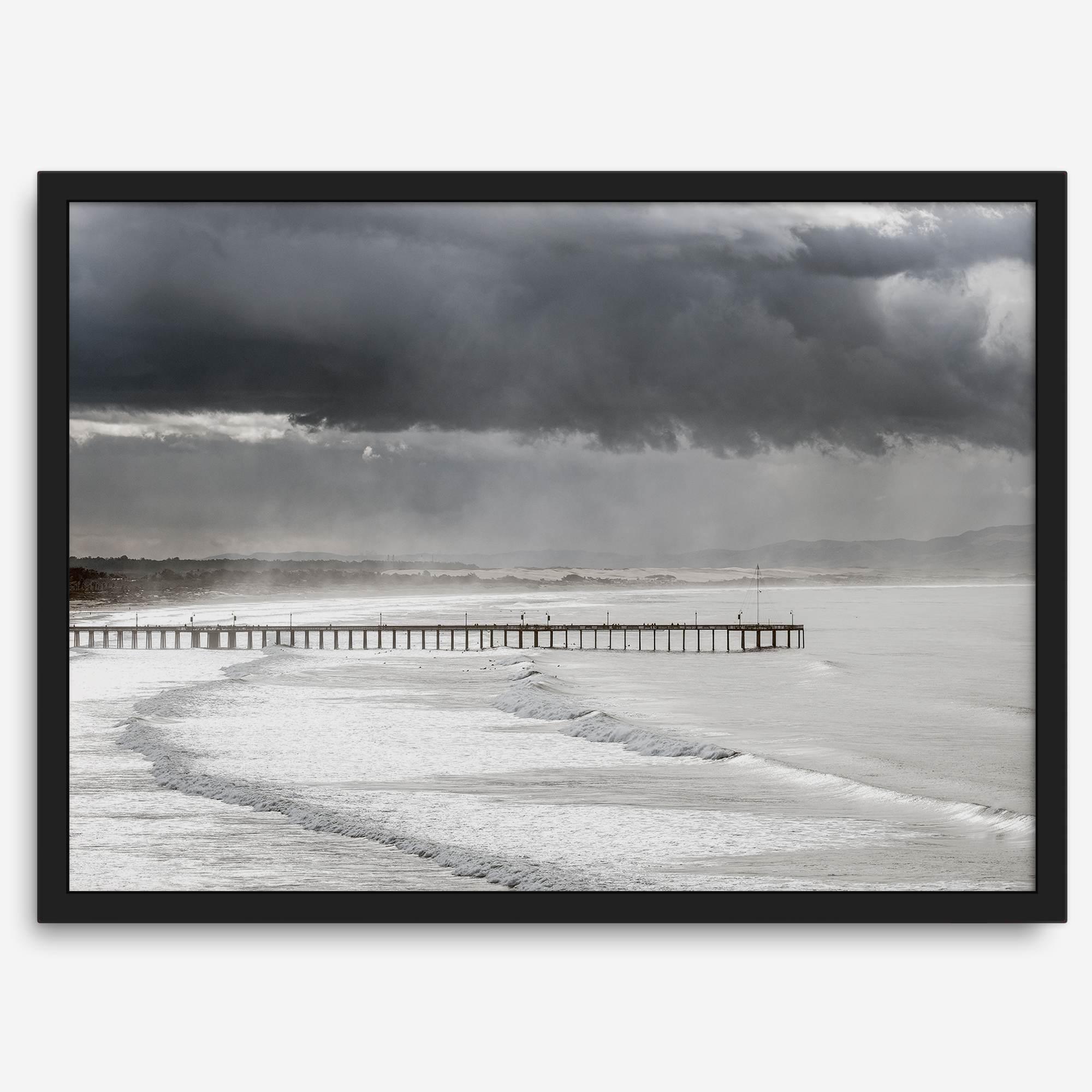 This fine art ocean photography print was taken in Pismo Beach, California, and showcases a distant view of an ocean pier and stormy clouds. Looking long and hard, you can see tens of surfers around the pier.