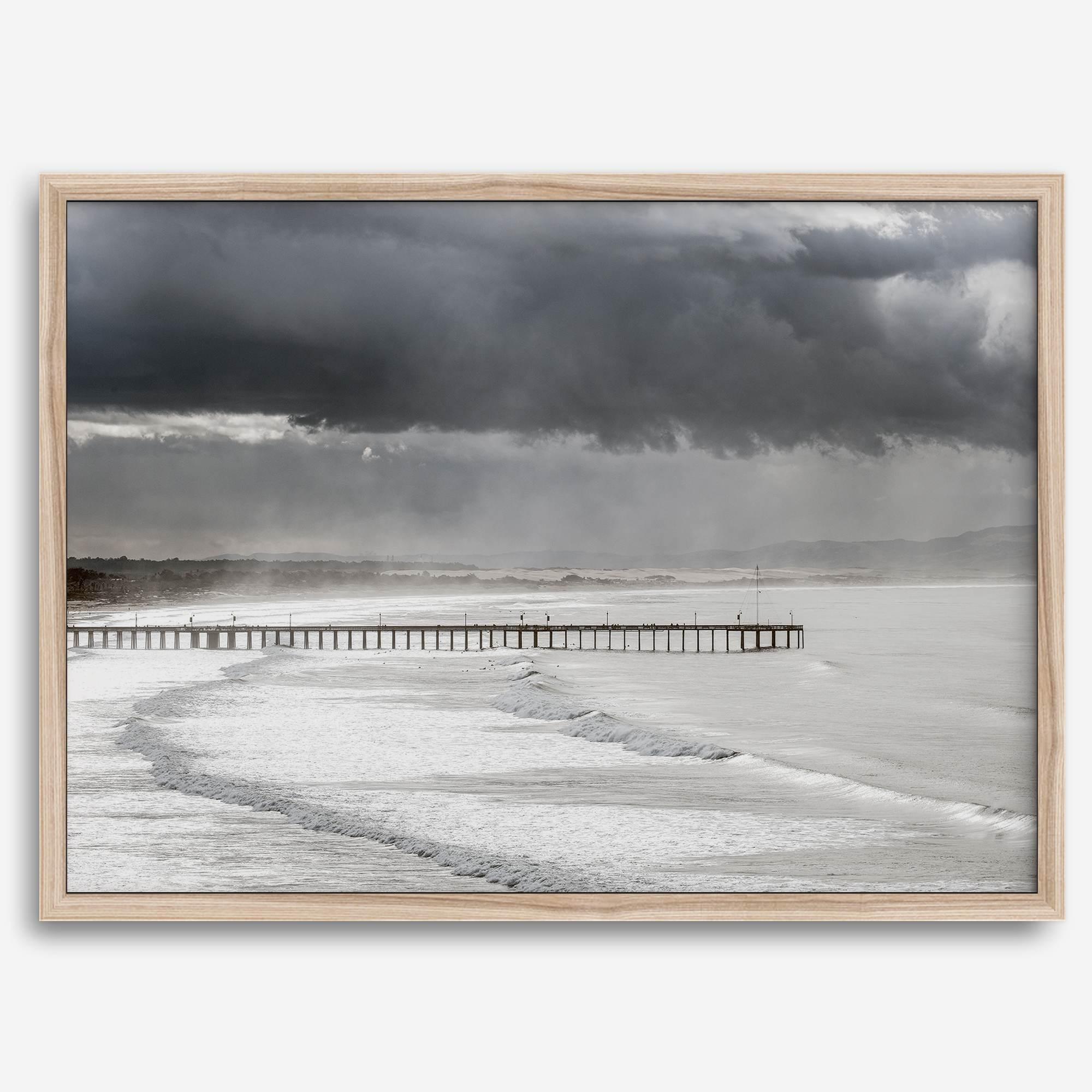 This fine art ocean photography print was taken in Pismo Beach, California, and showcases a distant view of an ocean pier and stormy clouds. Looking long and hard, you can see tens of surfers around the pier.