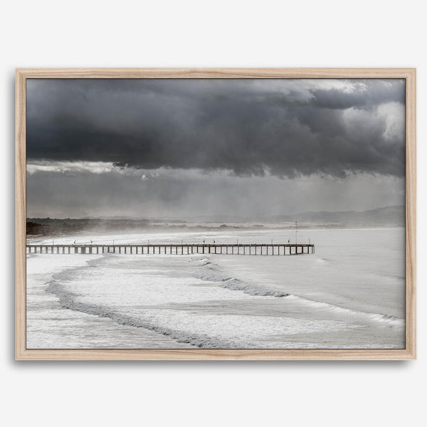 This fine art ocean photography print was taken in Pismo Beach, California, and showcases a distant view of an ocean pier and stormy clouds. Looking long and hard, you can see tens of surfers around the pier.
