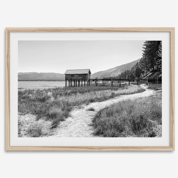 A fine art black and white vintage style framed photography print featuring a rustic old boathouse nested on a Lake Tahoe beach.