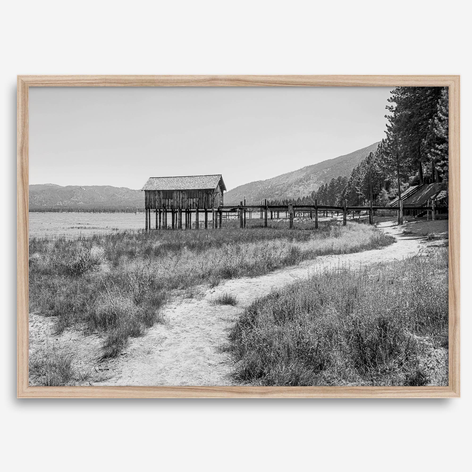 A fine art black and white vintage style framed photography print featuring a rustic old boathouse nested on a Lake Tahoe beach.