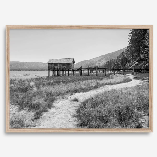 A fine art black and white vintage style framed photography print featuring a rustic old boathouse nested on a Lake Tahoe beach.