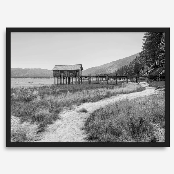 A fine art black and white vintage style framed photography print featuring a rustic old boathouse nested on a Lake Tahoe beach.
