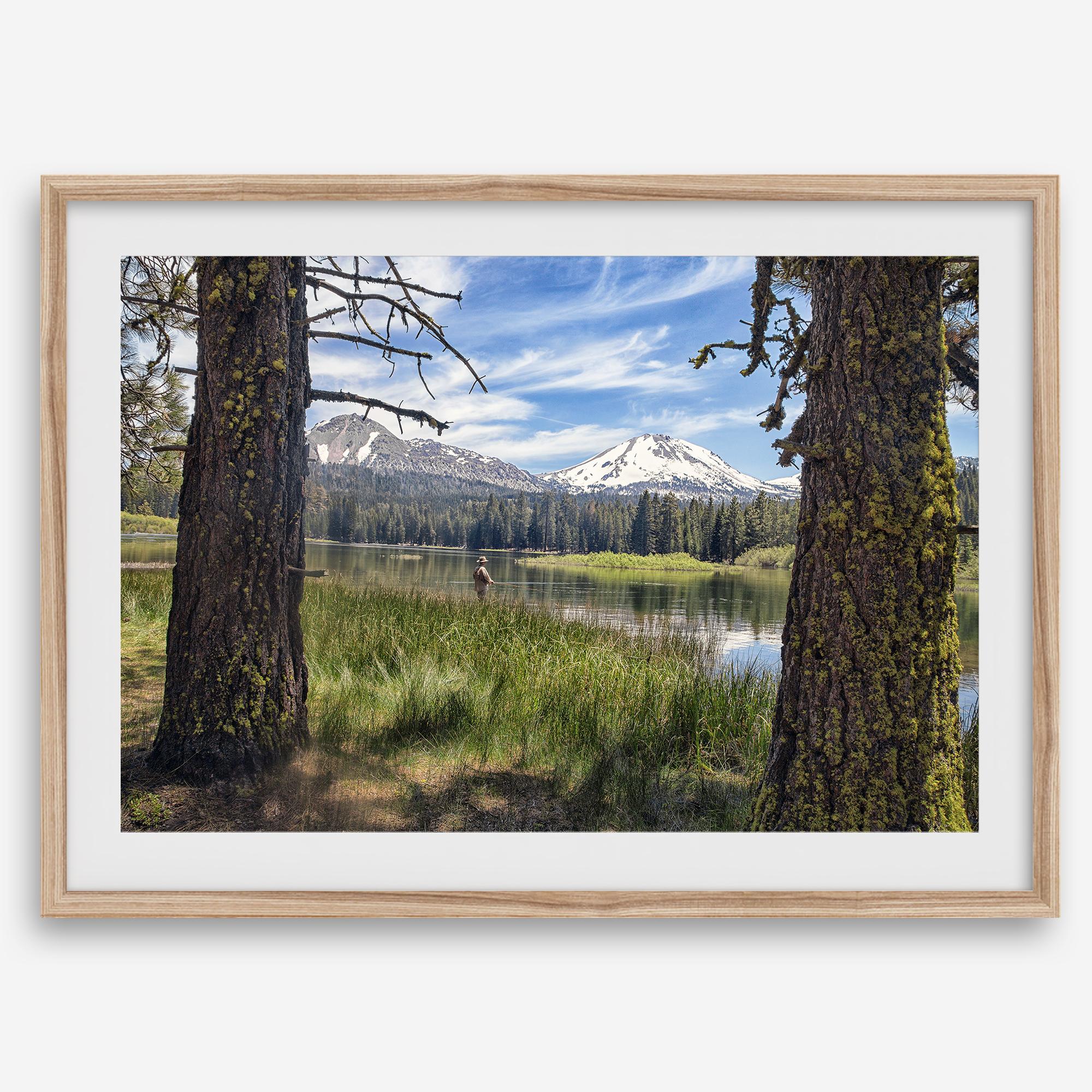 Fisherman at Manzanita Lake beneath snowy mountains and pine forest, photographed as peaceful fishing wall art.