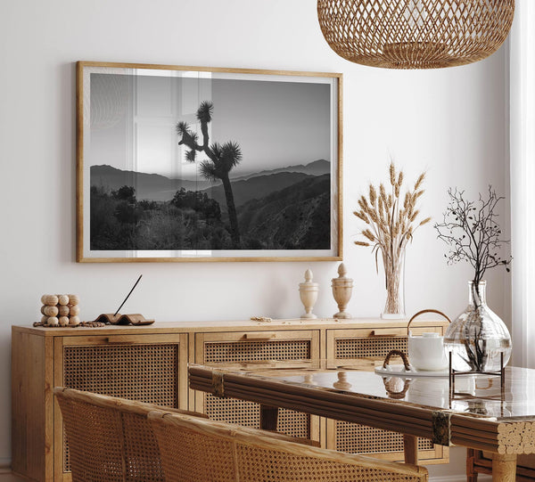 A fine art black and white desert print of a lone Joshua Tree against a sunset with the layered desert mountains behind it.