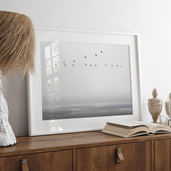 Minimalist seascape fine art print – a flock of birds in flight over a calm, misty ocean with a soft, gray horizon.