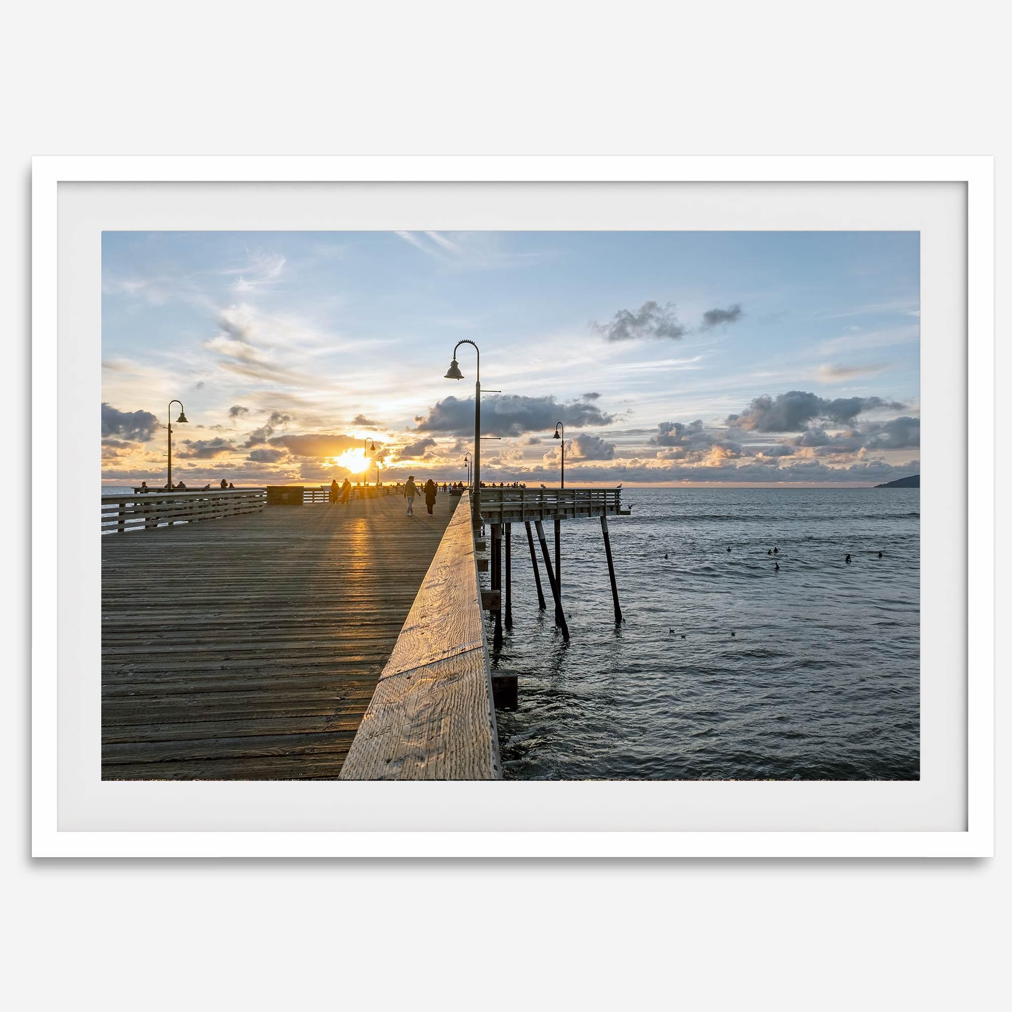 A fine art ocean sunset print featuring Pismo Beach Pier in Sunset with the surfers in the ocean and people enjoying a stroll on the pier.