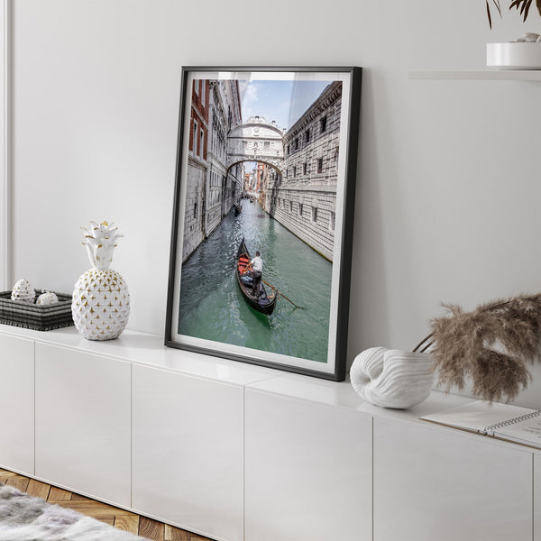 Venice canal photography print featuring a gondola approaching the Bridge of Sighs between historic stone buildings in Venice, Italy.
