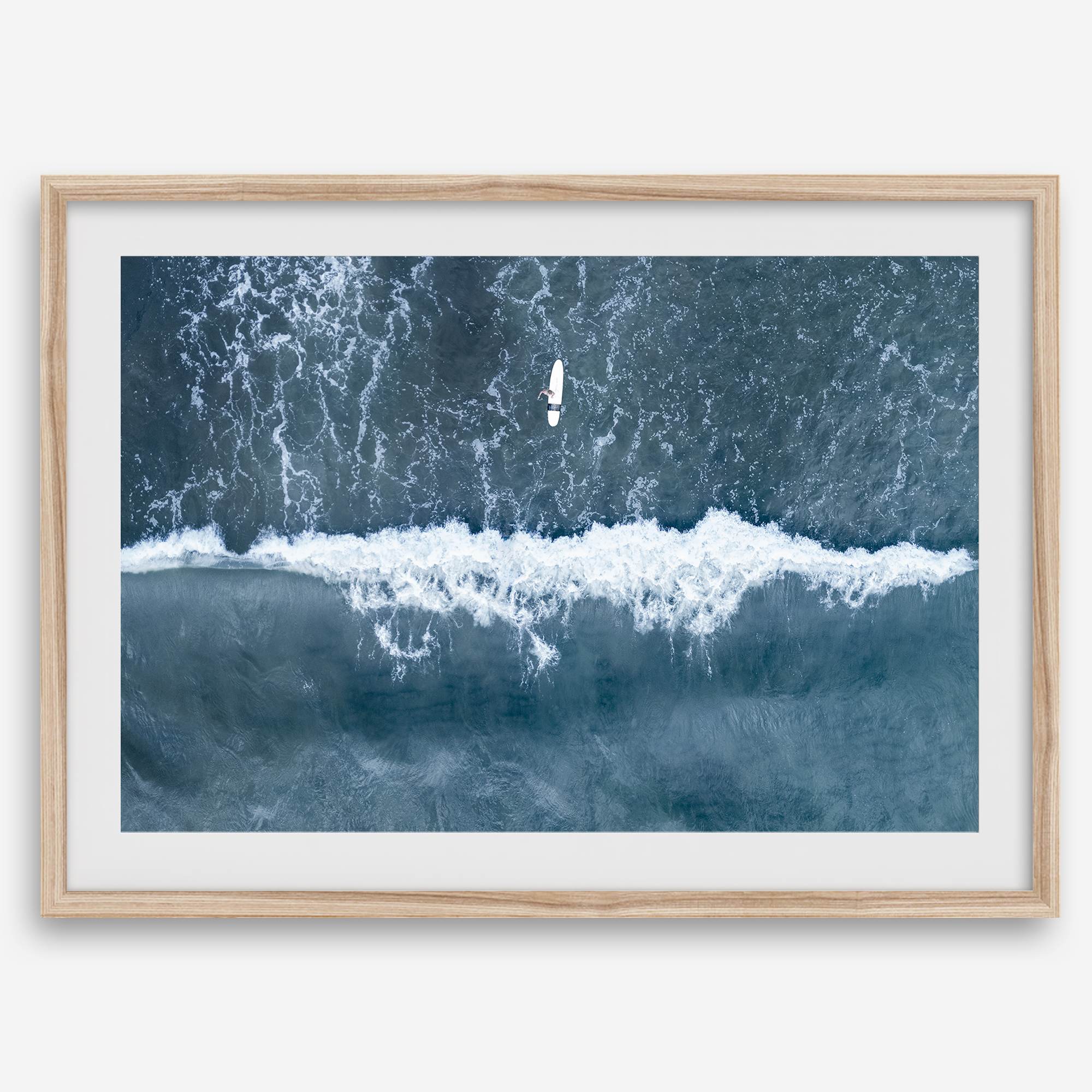 Aerial ocean wall art featuring a lone surfer and a breaking white wave in the deep blue waters of Hanalei Bay.