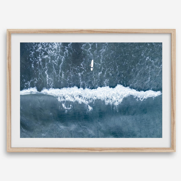 Aerial ocean wall art featuring a lone surfer and a breaking white wave in the deep blue waters of Hanalei Bay.