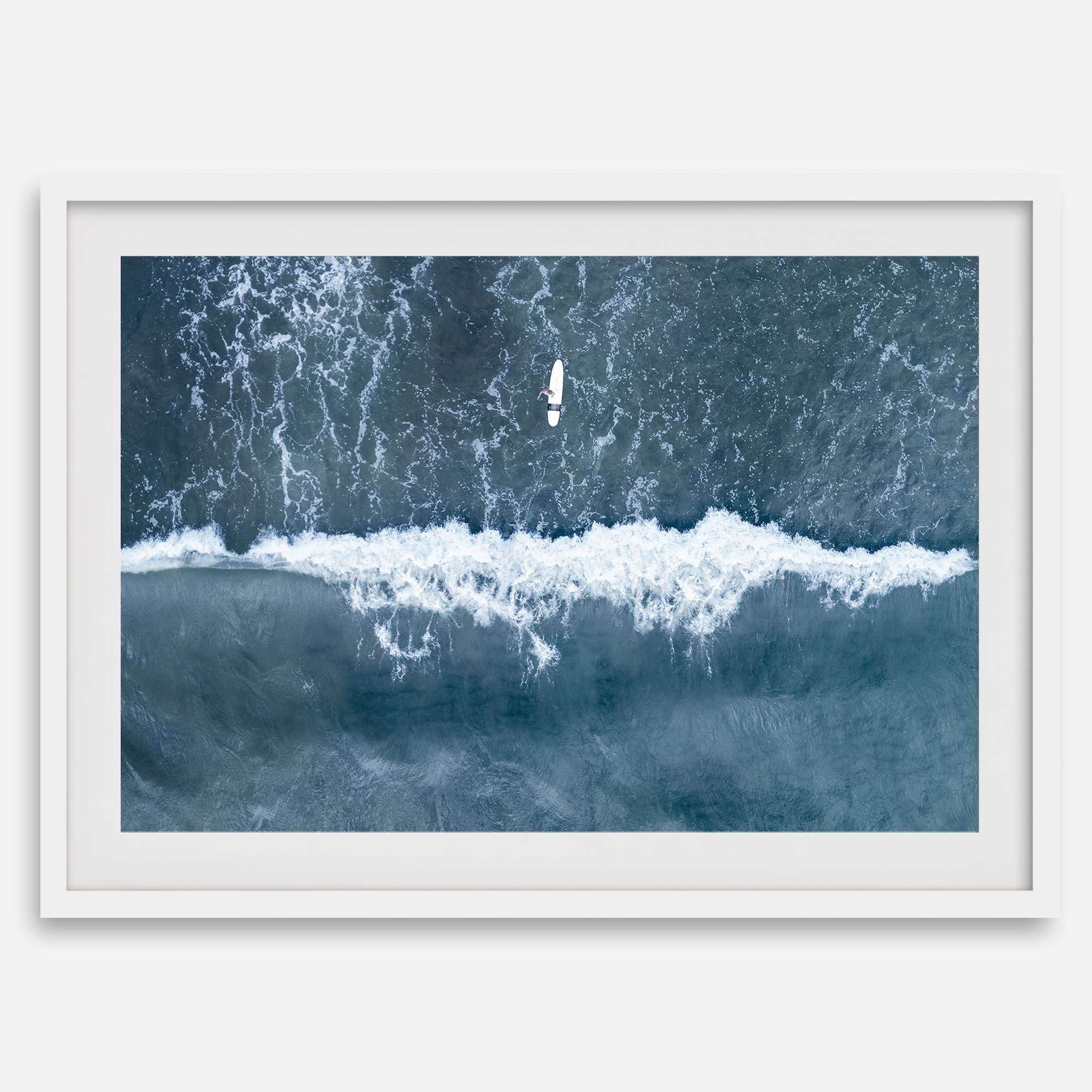 Aerial ocean wall art featuring a lone surfer and a breaking white wave in the deep blue waters of Hanalei Bay.