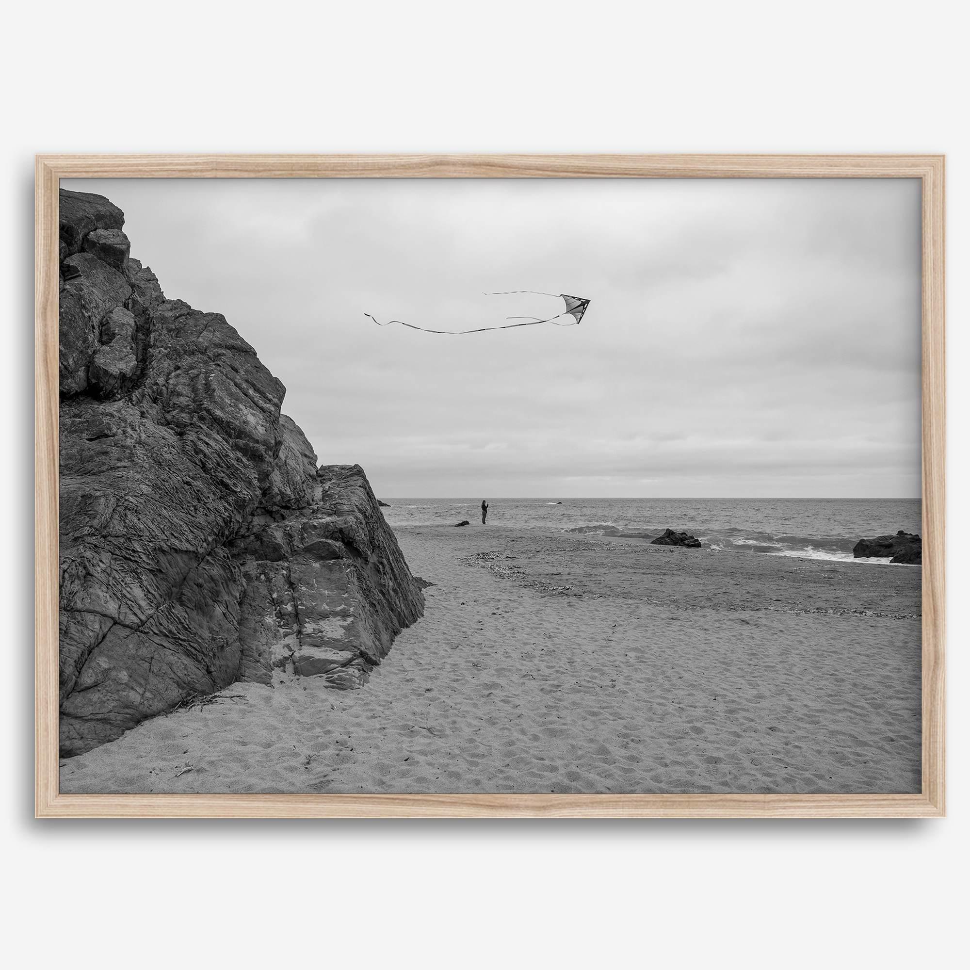 Black-and-white framed photo of a kite flying over a quiet California beach with rocky cliffs and an overcast sky.