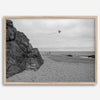 Black-and-white framed photo of a kite flying over a quiet California beach with rocky cliffs and an overcast sky.