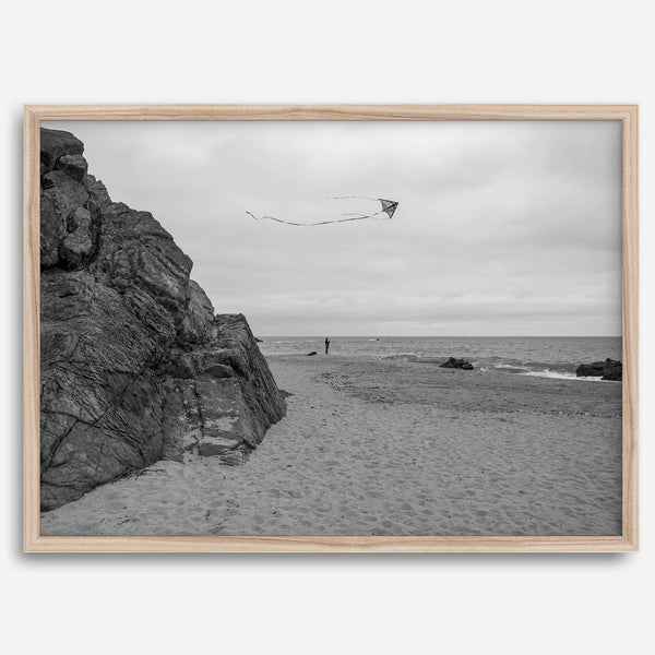 Black-and-white framed photo of a kite flying over a quiet California beach with rocky cliffs and an overcast sky.