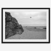 Black-and-white framed photo of a kite flying over a quiet California beach with rocky cliffs and an overcast sky.
