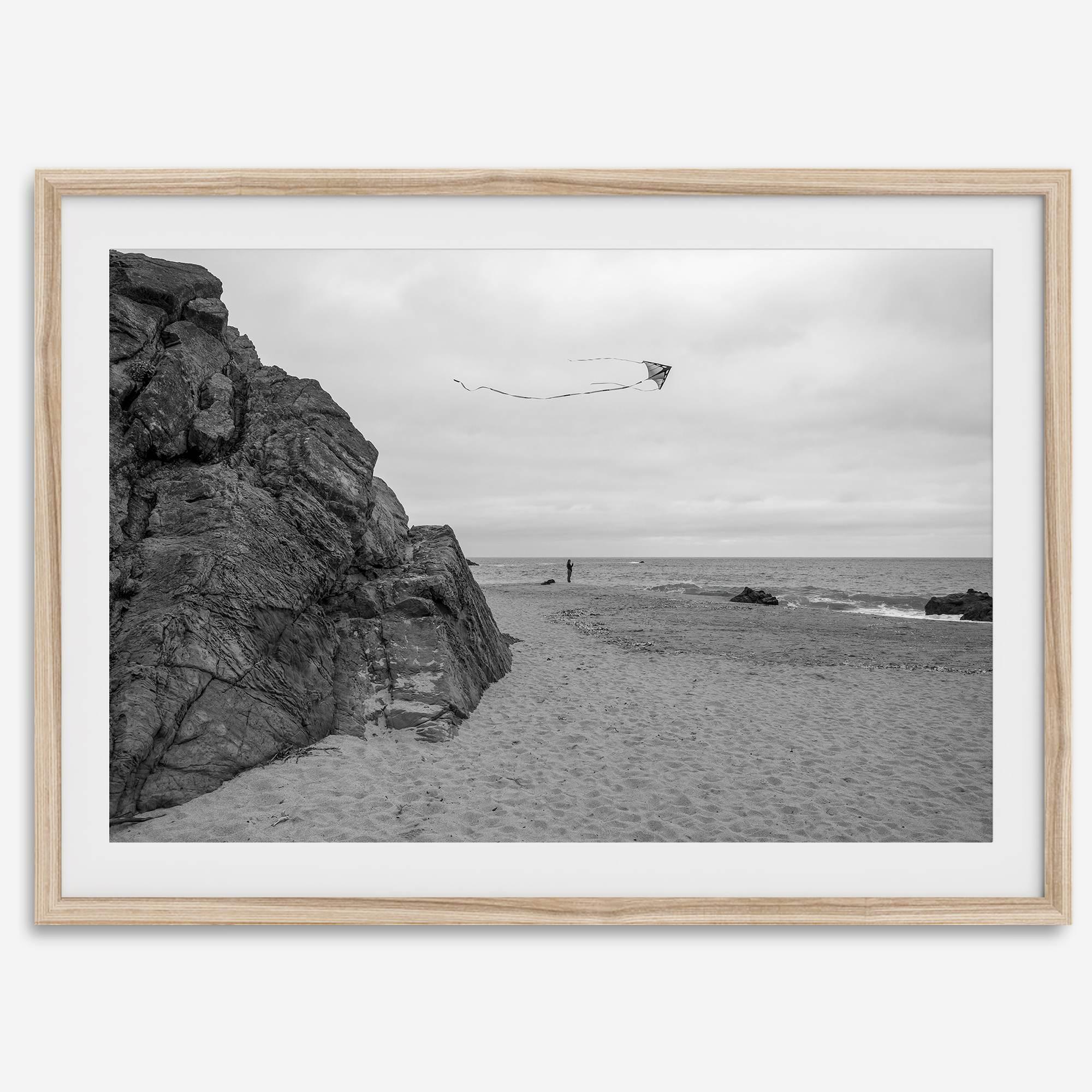 Black-and-white framed photo of a kite flying over a quiet California beach with rocky cliffs and an overcast sky.