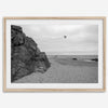 Black-and-white framed photo of a kite flying over a quiet California beach with rocky cliffs and an overcast sky.