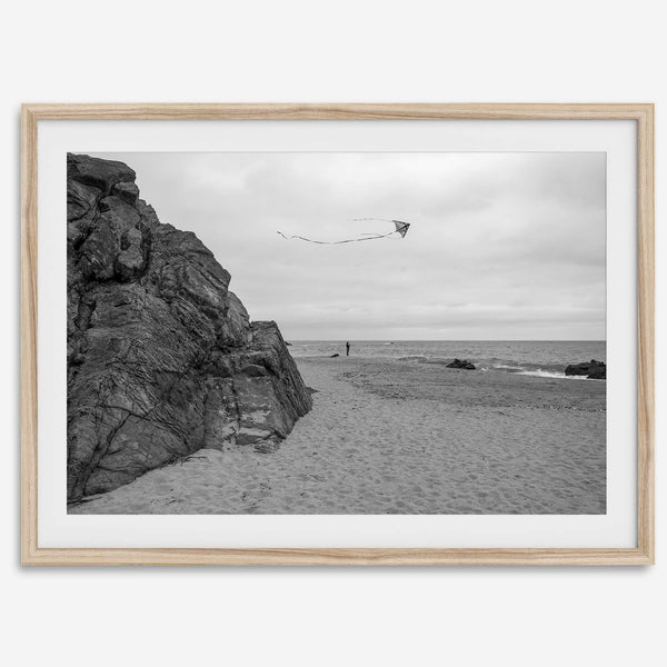 Black-and-white framed photo of a kite flying over a quiet California beach with rocky cliffs and an overcast sky.