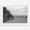 Black-and-white framed photo of a kite flying over a quiet California beach with rocky cliffs and an overcast sky.