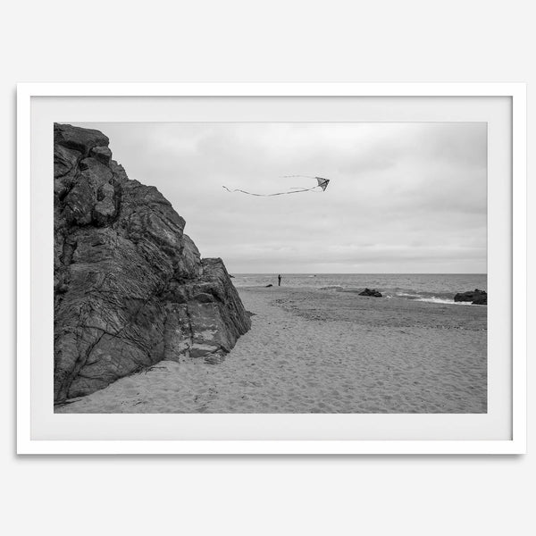 Black-and-white framed photo of a kite flying over a quiet California beach with rocky cliffs and an overcast sky.